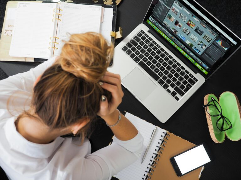 Overhead view of a stressed woman working at a desk with a laptop, phone, and notebooks trying to cope with managing stress